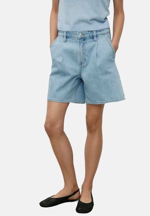 Person iført lyseblå denimshorts, lyseblå ærmeløs top, med hænderne i lommerne og sorte flade sko.