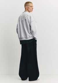 Grå sweatshirt med stort sort motiv på ryggen, kombineret med mørk denim bukser med vide ben og hvide sneakers. Simpelt, afslappet design.