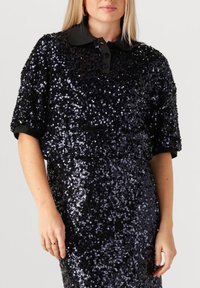 Vrouw in een zwart pailletten kortemouwen jurk met een zwarte kraag en knopenlijst, met haar hand langs haar zijde.