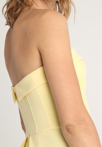 Vestido amarillo sin mangas con un corsé ajustado, que presenta costuras verticales y un detalle de lazo en la parte posterior. Tejido suave y texturizado.