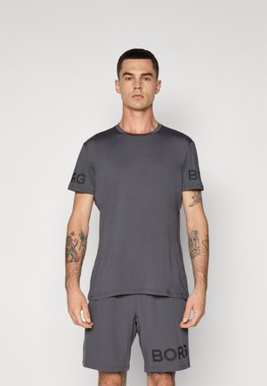 Mann mit Tattoos, der ein passendes dunkelgraues Sport-T-Shirt und Shorts mit dem Text "BORG" trägt, steht vor einem schlichten hellen Hintergrund.