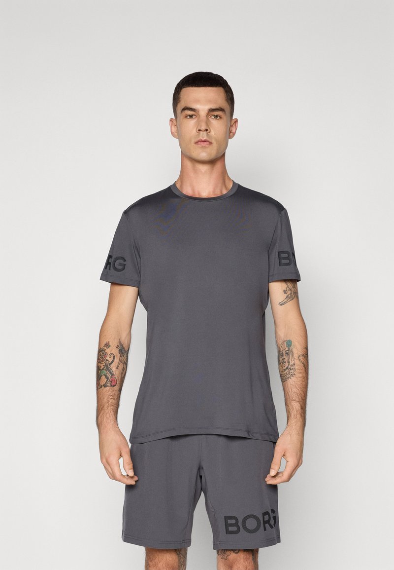 Mann mit Tattoos, der ein passendes dunkelgraues Sport-T-Shirt und Shorts mit dem Text "BORG" trägt, steht vor einem schlichten hellen Hintergrund.