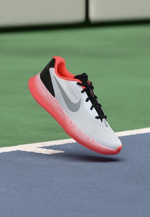 Hvid og pink Nike løbesko med sorte snørebånd, der svæver over en blå og grøn sportsbane.