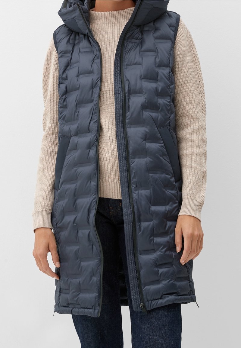 Personne portant un long gilet matelassé sans manches bleu foncé sur un pull en maille beige et un pantalon foncé, debout les bras détendus.