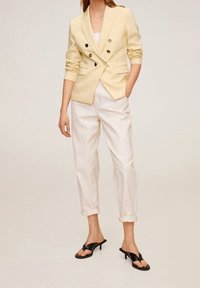 Donna con blazer doppiopetto giallo chiaro, pantaloni bianchi arrotolati e sandali neri con tacco, in piedi davanti a uno sfondo neutro.