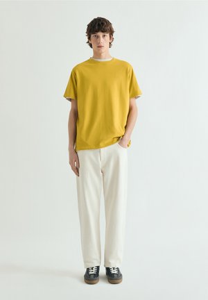 Joven de pie contra un fondo blanco, vestido con una camiseta amarilla mostaza, pantalones blanco hueso y zapatillas negras con cordones blancos.