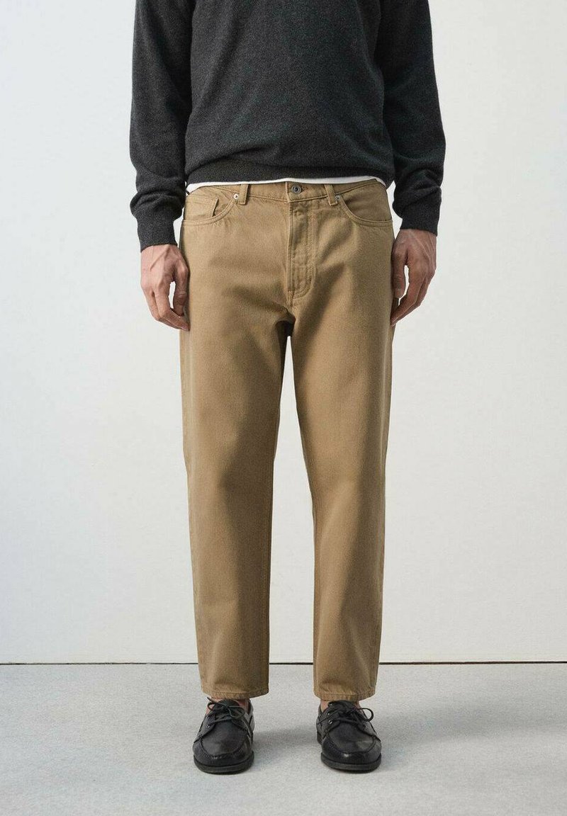 Homme portant un pantalon beige à jambes droites, des chaussures noires et un pull à manches longues foncé, debout sur un sol gris clair contre un mur blanc.