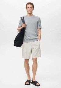 Gray ribbed short-sleeve shirt, light beige shorts, and black sandals. Model carries a black tote bag, standing against a neutral background.