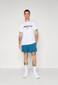 Vit T-shirt med texten "Just Do It" i marinblått och blå shorts. Modellen bär vita sneakers och strumpor, vilket visar upp en ren atletisk stil.