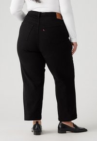 Black high-waisted jeans with a tapered leg, featuring back pockets and a leather patch on the waist. Worn with black flat shoes.