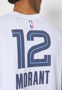 T-shirt en coton blanc avec le numéro "12" en marine, doté de détails bleu clair, et "MORANT" imprimé en marine en dessous. Logo NBA au niveau du col.