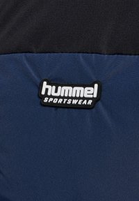 Gros plan sur une section de tissu bleu marine et noir avec un patch logo "hummel SPORTSWEAR" cousu dessus.