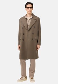 Abrigo marrón de doble botonadura con grandes botones negros, solapas peak y un corte relajado, usado sobre una camisa beige y pantalones beige claros.