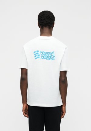 Person with short curly hair wears a white T-shirt with repeated blue "KARL LAGERFELD" text on the upper back, standing against a plain background.