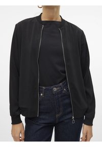 Blouson bomber noir avec une texture lisse, des poignets ajustés et une fermeture éclair argentée. Porté sur un haut noir, associé à un jean foncé taille haute.