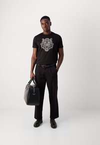 Zwart katoenen T-shirt met een witte tijgerafbeelding, gecombineerd met zwarte broeken en zwarte leren laarzen, en een zwarte leren tas in hand.