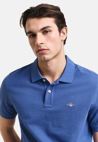 Mladý muž s tmavými vlasy, který nosí modré polo tričko s krátkým rukávem a malým vyšitým logem na hrudi.