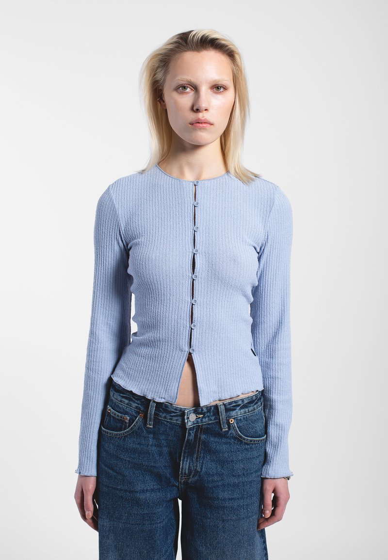 Ljusblå ribbad långärmad tröja med rund halsringning, framknäppning och något kortare nederkant. Bärs tillsammans med mörkblå högmidjade jeans.