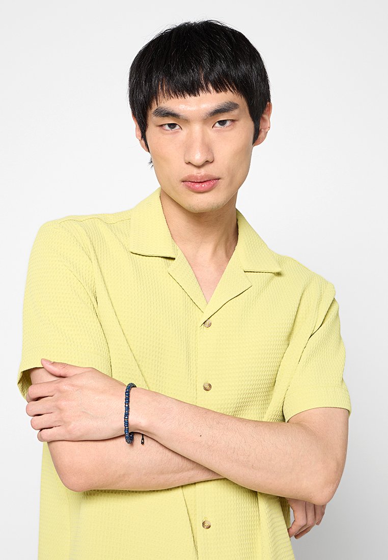 Helles gelbes Kurzarmhemd mit texturiertem Muster und Knöpfen. Das Model trägt ein dunkelblaues Perlenarmband am Handgelenk, mit verschränkten Armen.