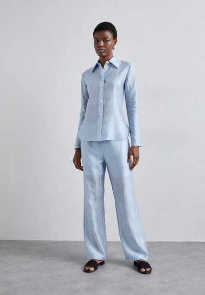 Chemise en lin bleu clair à manches longues et fermeture à boutons, associée à un pantalon en lin bleu clair assorti, et des sandales glissantes noires.