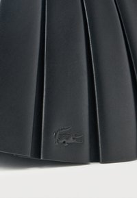 Μαύρη πλισέ δερμάτινη φούστα με ανάγλυφο λογότυπο κροκοδείλου κοντά στο τελείωμα, σε λευκό φόντο.