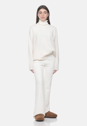 Woman with long dark hair wearing a white turtleneck sweater, white flared pants, and brown fuzzy slippers, standing against a light background.