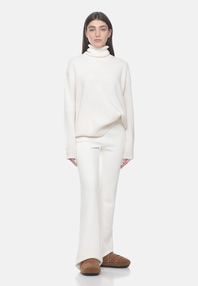 Femme aux longs cheveux foncés portant un pull à col roulé blanc, un pantalon évasé blanc et des chaussons marron en peluche, debout devant un fond clair.