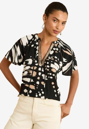 Donna che indossa una blusa a maniche corte con un motivo astratto in bianco e nero e pantaloni beige, in piedi con una mano su un fianco.