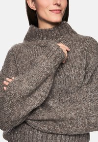 Pull en mailles épaisses dans un mélange de tons bruns et gris. Col montant, poignets côtelés et corps large. Texture douce avec une apparence confortable.