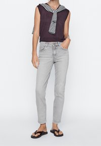 Jean gris clair avec une coupe ajustée et cinq poches, associé à un top sans manches violet foncé et un pull rayé noir et blanc noué autour du cou.