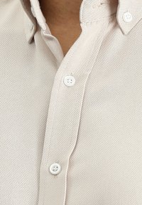 Camicia beige con bottoni, realizzata in un tessuto testurizzato, dotata di collo, due bottoni bianchi e dettagli di cucitura visibili lungo la pattina.
