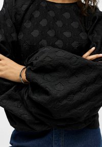 Persoon met gekruiste armen, gekleed in een zwarte gestructureerde blouse met opbollende mouwen en blauwe jeans, vergezeld van een zilveren armband.