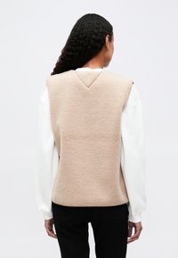 Vest din fleece bej, cu o cusătură în formă de V pe spate, design fără mâneci și material texturat. Purtați peste o cămașă albă cu mânecă lungă.