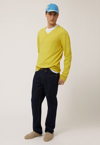 Geel gebreid trui met een V-hals, donkerblauwe denim jeans en beige instap schoenen. De pet heeft blauw en witte strepen.