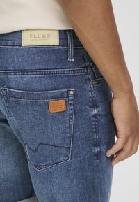 Jeans en denim de couleur foncée, avec un patch en cuir étiqueté "BLEND" sur la taille, et deux poches arrière comportant des détails de couture subtils.