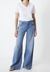 Mujer vistiendo una camiseta blanca de cuello en V y manga corta, pantalones vaqueros de cintura alta y pierna ancha azules, y zapatos dorados de punta, de pie contra un fondo blanco.