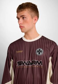 Braun gestreiftes Langarmtrikot mit cremefarbenen Akzenten, mit einem Logo auf der Brust und dem Schriftzug "SWARM" in fetter Schrift. Hergestellt aus synthetischem Stoff.