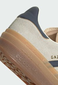 Adidas Gazelle sneaker with a light beige suede upper, dark blue leather accents, and a translucent rubber sole featuring embossed branding.