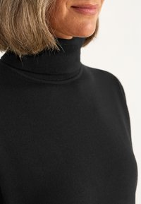 Frau mit schulterlangem, hellbraunem Haar, die einen schwarzen Rollkragenpullover vor einem weißen Hintergrund trägt.