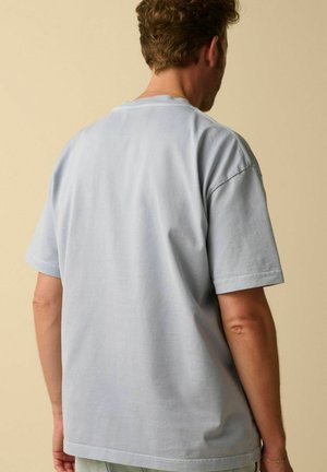 Man with curly hair wearing a light blue oversized short-sleeve t-shirt, shown from the back against a beige background.