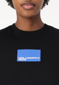 Črn bombažni majica s kratkimi rokavi, na katerem je modra pravokotna grafika z belim napisom "KARL LAGERFELD JEANS" v centru na prsih.