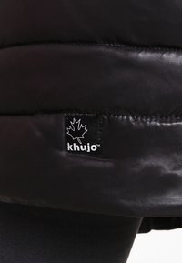 Sort pufferjakke lavet af skinnende stof. Har et kviltet design og et sort mærke med et hvidt ahornblad og mærkenavnet "khujo."