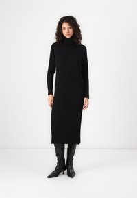 Black ribbed knit dress with a high collar, long sleeves, and side slits, worn with knee-high black pointed-toe boots.