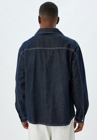 Mörk denimjacka med lös passform, som har en spetsig krage, knappstängning och kontrasterande vit söm. Bakvyn visar minimal detaljering.