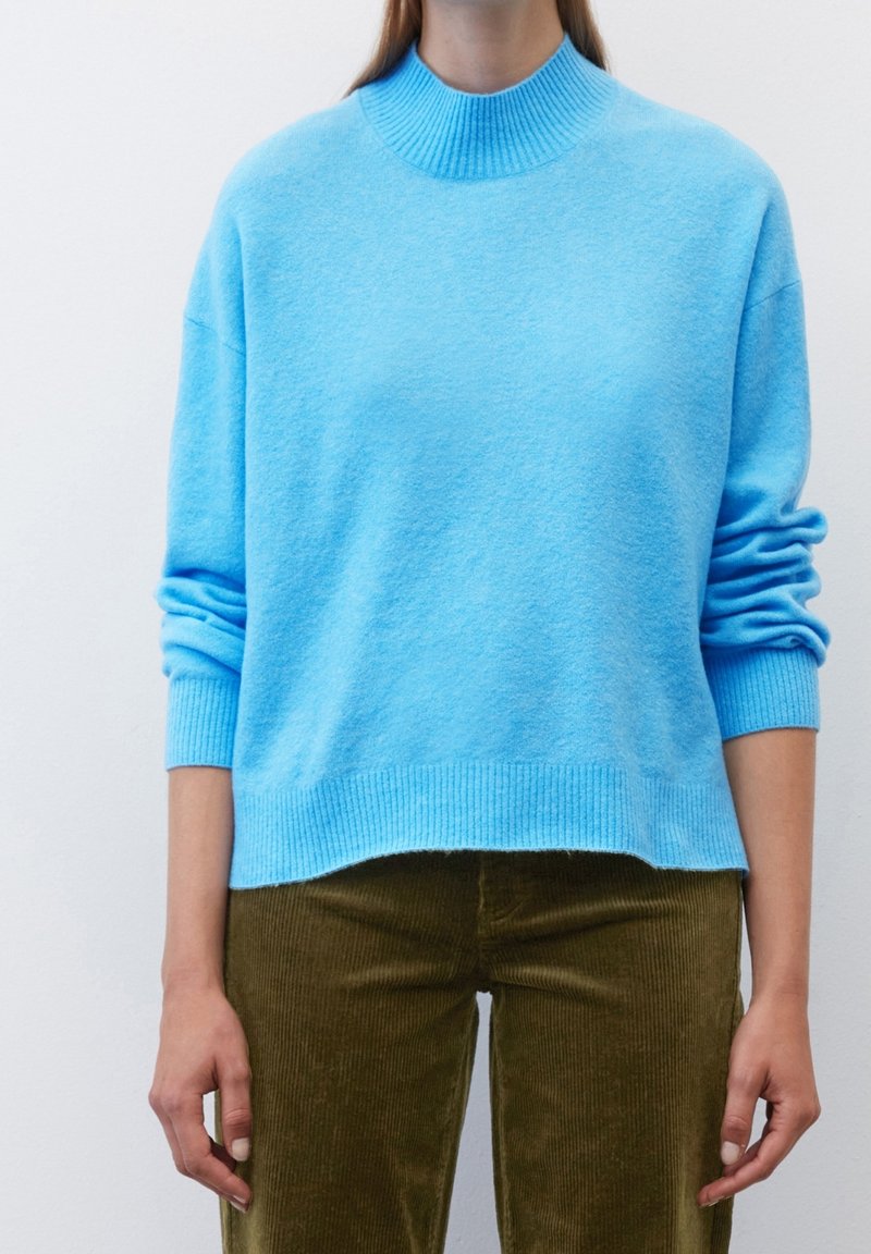 Pull en maille bleu clair avec un col montant côtelé et des manches longues. Présente une coupe décontractée et des poignets et un ourlet côtelés. Accent sur la texture.