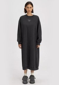 Oversized zwarte katoen jurk met lange mouwen, ronde hals en zijsplitten, met minimalistische witte tekst op de borst.