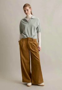 Zaļa adīta blūze ar polo apkakli, saskaņota ar platām brūnām kordurojām biksēm ar pleciem un augstu jostas vietu. Modelis valkā vieglu apavu.