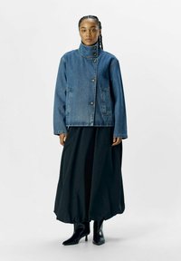 Veste en denim avec un col haut, des boutons à l'avant et des poches latérales, assortie d'une jupe noire fluide et de bottes noires à bout pointu.