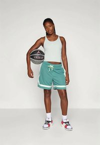 Vihreä tankkitoppi, teal-väriset shortsit valkoisilla yksityiskohdilla, ja mustat sekä vaaleanpunaiset lenkkarit. Pitelee mustaa koripalloa, jossa on vaaleanpunainen logo.