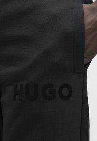 Close-up van zwarte broek met het ingegraveerde "HUGO"-logo op het dijbeen en een hand in de zak, tegen een lichte achtergrond.